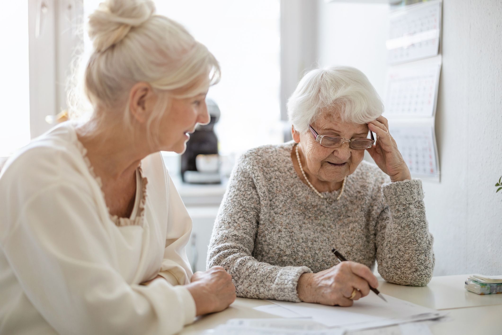 Two elderly women sitting at a table, one writing on a piece of paper while the other watches and smiles, with a calendar visible on the wall behind them.