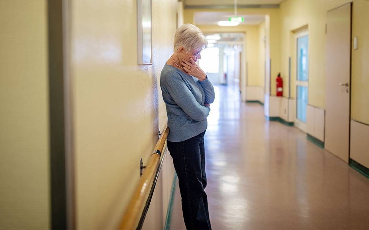 Elderly woman with short white hair, wearing a blue sweater and black pants, stands leaning against a wall in a long, empty hallway with a hand on her chin, appearing deep in thought.