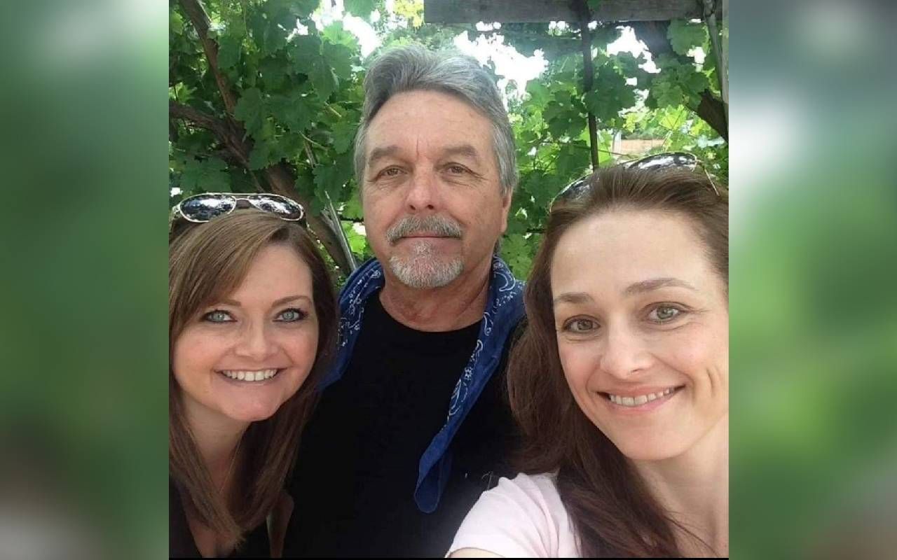 Three people smiling for a selfie outdoors with green leafy plants in the background. Two women, one on the left and one on the right, flank a man in the middle. The man has gray hair and a gray beard, and is wearing a black shirt with a blue bandana around his neck. Both women have long hair and are wearing sunglasses on their heads.