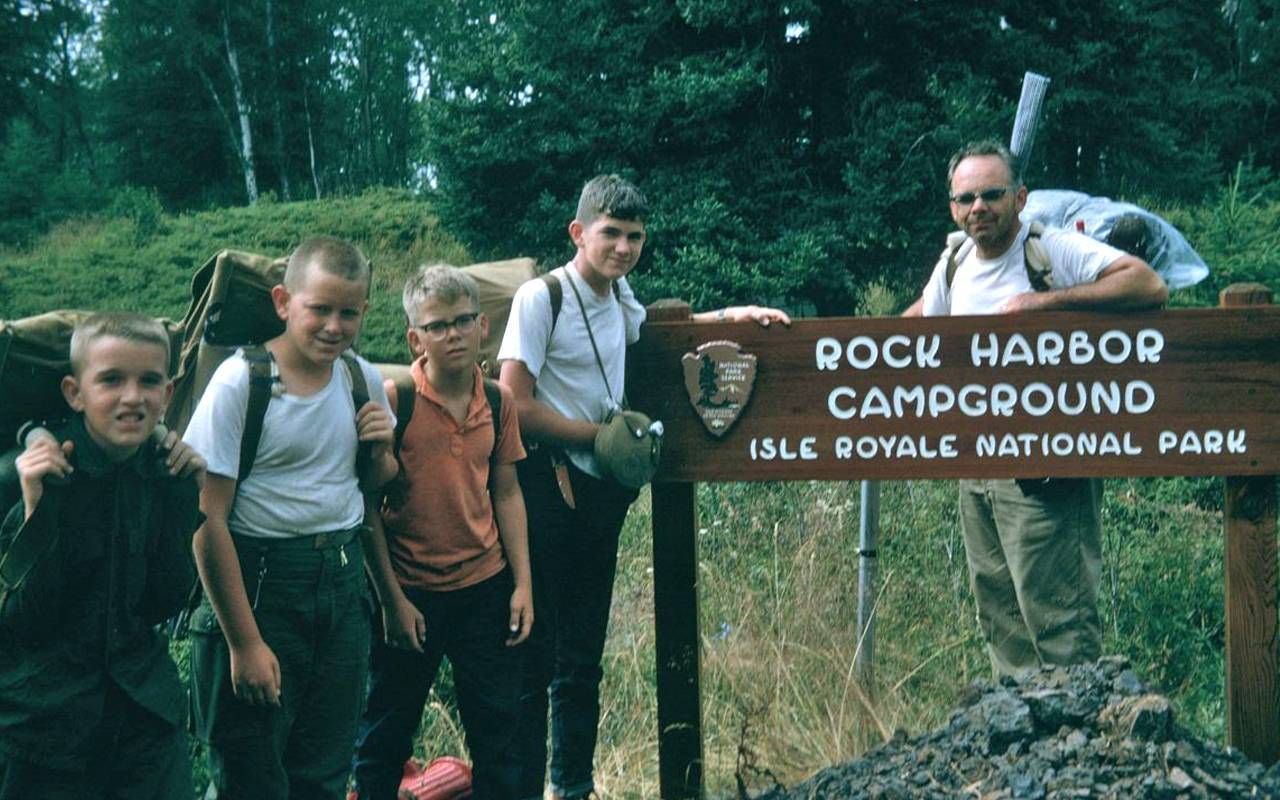 A group of five boys and a man pose in front of a wooden sign reading "Rock Harbor Campground, Isle Royale National Park." The boys, wearing casual hiking attire and backpacks, display a range of expressions, from smiles to serious looks. The man stands to the right, wearing sunglasses and a backpack. The background features trees and greenery typical of a wilderness area.