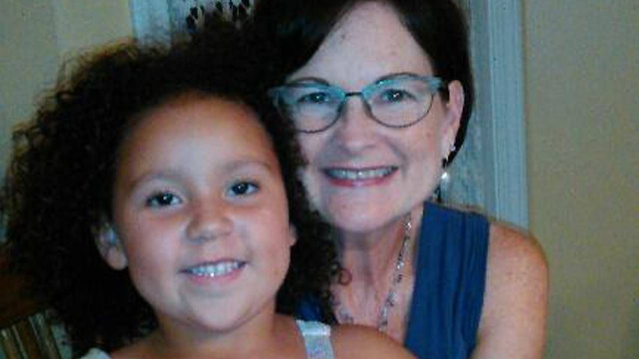 Two smiling people, a young girl with curly hair in the foreground and an older woman with short dark hair and glasses behind her, both looking at the camera.